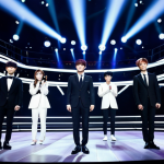 A professional group of diverse K-Pop artists performing on a grand, illuminated stage. They are fully clothed in modest, stylish, and professional stage attire, such as tailored, full-coverage suits or elegant, modern ensembles. The vibrant background showcases a futuristic concert arena with dynamic stage lighting and a blurred, diverse global audience, emphasizing the genre's international reach. High-quality professional photography, perfect anatomy, correct proportions, well-formed hands, proper finger count, natural body proportions, natural pose, crisp focus, cinematic lighting. Safe for work, appropriate content, family-friendly.